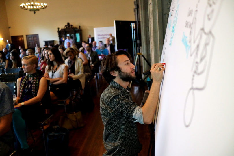 Graphic Recorder scribing at a board in front of an audience of conference attendees.